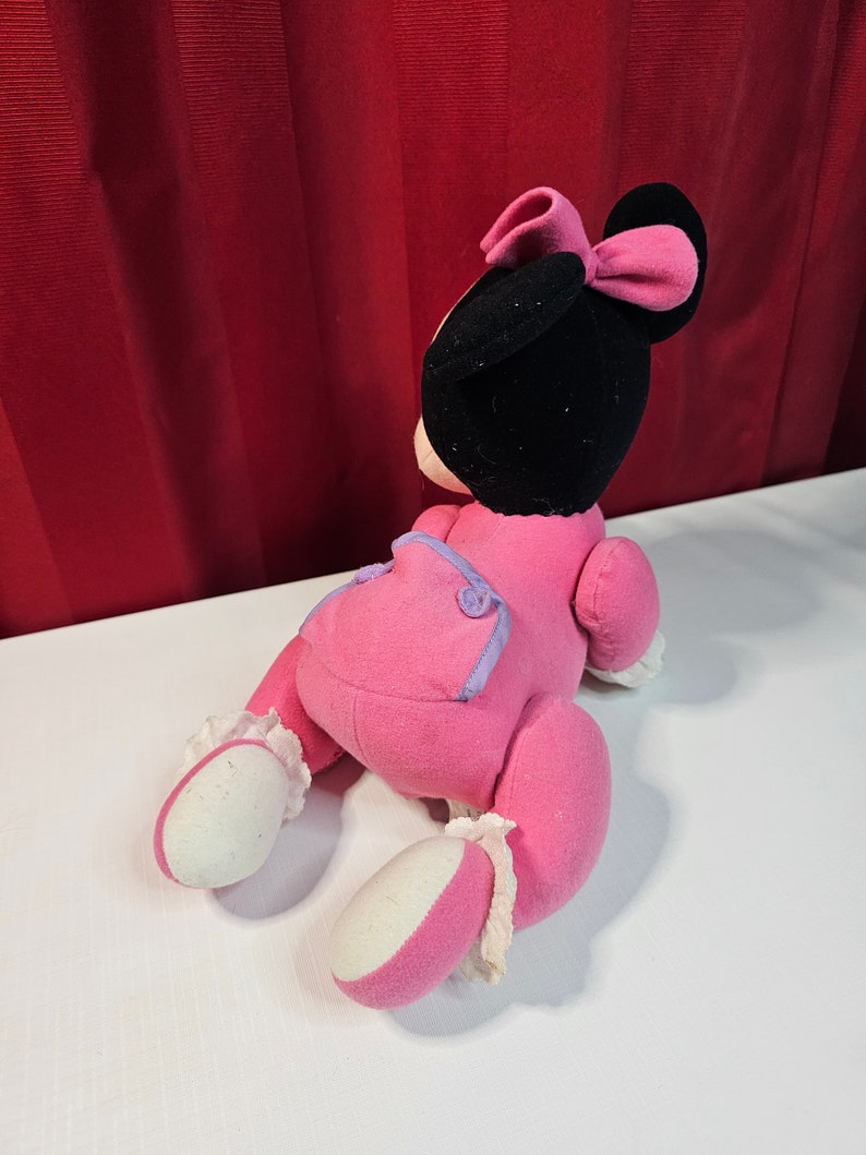 K&ouml;nnte beinhalten: Rosa Pl&uuml;schtier einer Comicmaus mit schwarzem Kopf und einer rosa Schleife. Das Spielzeug tr&auml;gt ein rosa-violettes Outfit mit wei&szlig;en Akzenten.