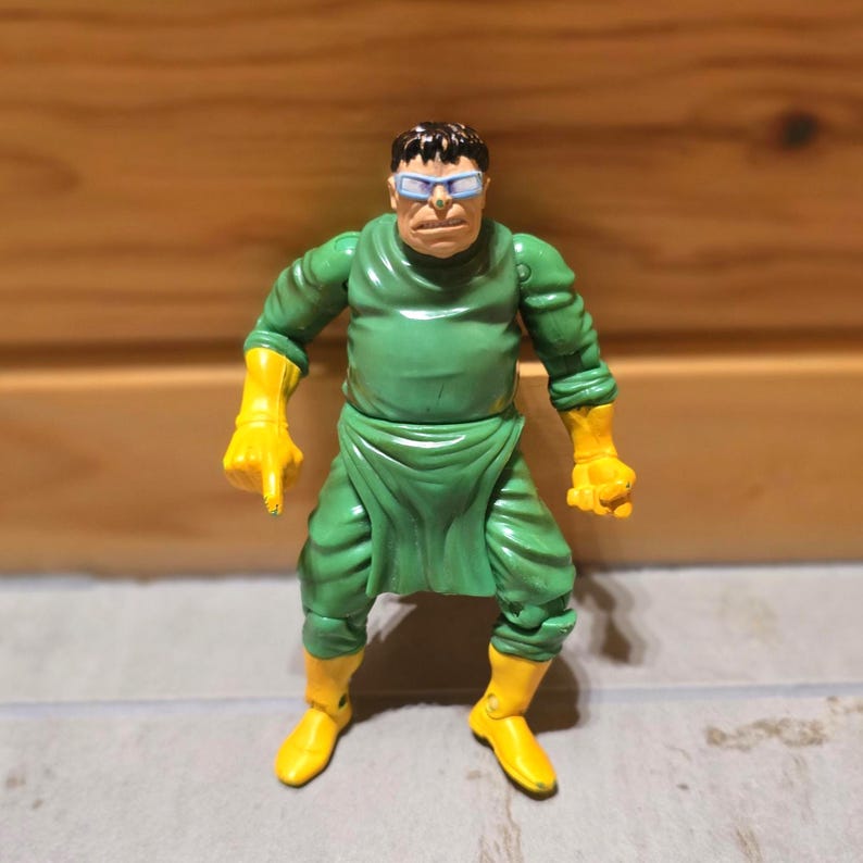 May include: A Dr. Octopus action figure. The figure is wearing a green outfit with yellow gloves and boots. The figure has a stern expression and is wearing blue-tinted goggles. The figure is standing in a neutral pose.