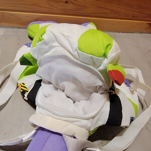 May include: A green, white, and black backpack with a zippered opening. The backpack is shaped like a cartoon character and has a white lining. The backpack has adjustable straps.