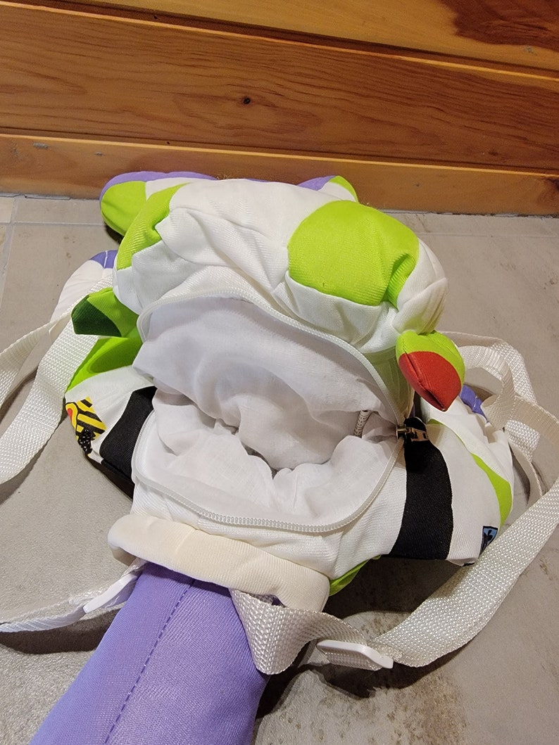 May include: A green, white, and black backpack with a zippered opening. The backpack is shaped like a cartoon character and has a white lining. The backpack has adjustable straps.