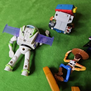 May include: A set of four plastic toy figures from the Toy Story franchise. The figures include Buzz Lightyear, Woody, and two other characters. The figures are all in different poses and are made of a variety of colours, including green, blue, red, yellow, and brown.