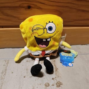 May include: Yellow plush toy of Spongebob Squarepants with a winking eye and a big smile. The toy is wearing a white shirt with a red tie and has black shoes. The toy is sitting on a light brown wooden surface. The tag says "Spongebob Squarepants".