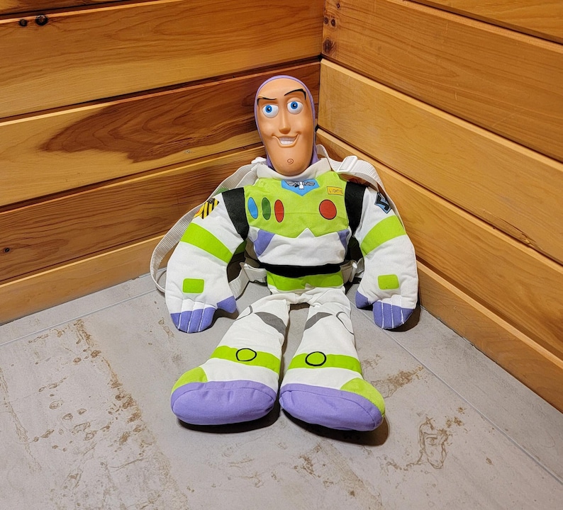 May include: A plush Buzz Lightyear backpack with a green and white spacesuit, purple boots, and a smiling face. The backpack is sitting on a wooden floor.
