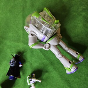 May include: A set of three plastic action figures from the Toy Story franchise. The figures are of Buzz Lightyear, a purple and black villain, and a smaller Buzz Lightyear figure. The larger Buzz Lightyear figure is in a space ranger suit with a clear plastic helmet and green and purple accents. The figures are on a green surface.