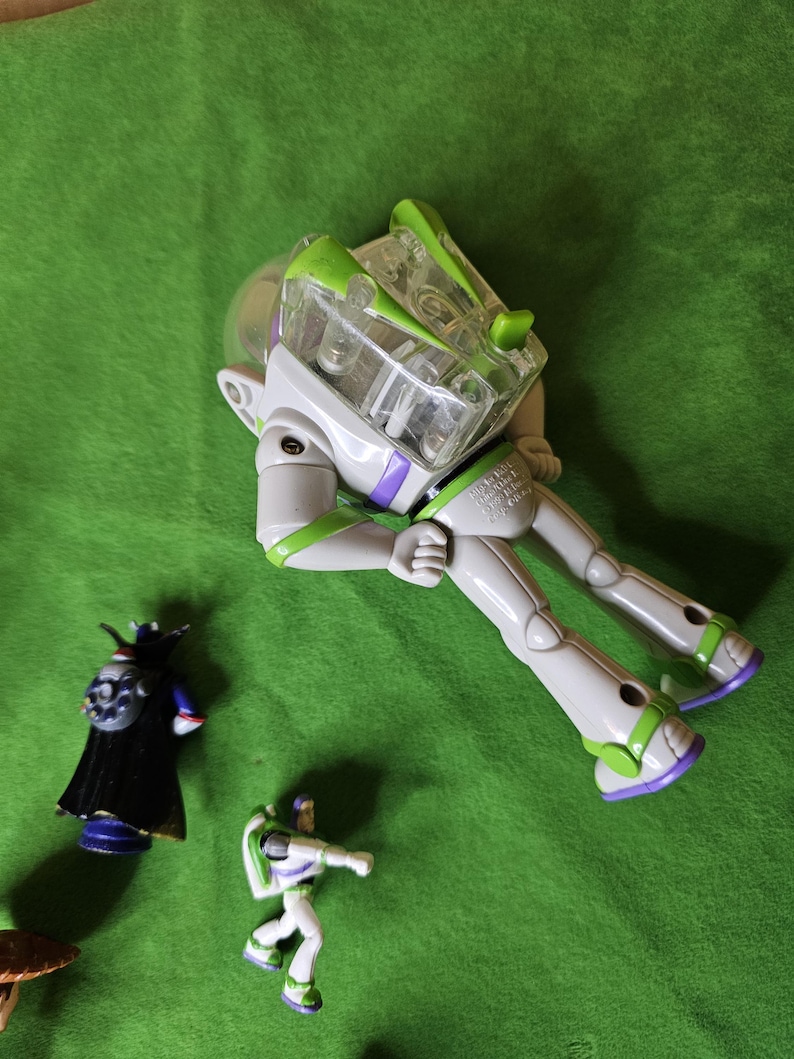 May include: A set of three plastic action figures from the Toy Story franchise. The figures are of Buzz Lightyear, a purple and black villain, and a smaller Buzz Lightyear figure. The larger Buzz Lightyear figure is in a space ranger suit with a clear plastic helmet and green and purple accents. The figures are on a green surface.