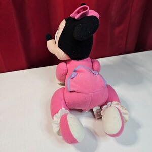 K&ouml;nnte beinhalten: Rosa Pl&uuml;schtier von Minnie Mouse in einem rosa und violetten Outfit, das auf einer wei&szlig;en Oberfl&auml;che kriecht.