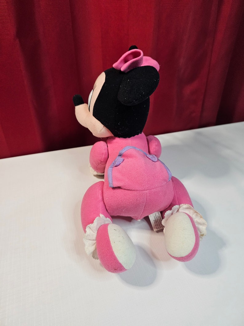 K&ouml;nnte beinhalten: Rosa Pl&uuml;schtier von Minnie Mouse in einem rosa und violetten Outfit, das auf einer wei&szlig;en Oberfl&auml;che kriecht.