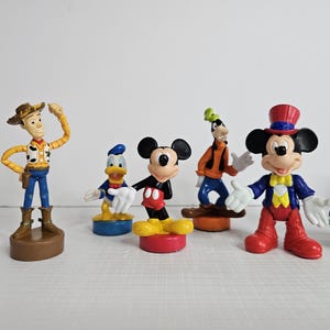 Op de afbeelding: Een set van zeven plastic Disney-figuren, waaronder Mickey Mouse, Minnie Mouse, Donald Duck, Goofy en Woody. De figuren zijn kleurrijk en gedetailleerd en zouden een geweldige aanvulling zijn op elke Disney-collectie.