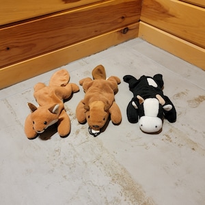 May include: Three plush animal toys: a fox, a beaver, and a cow. The fox is orange with white accents, the beaver is brown, and the cow is black and white. They are lying on a light gray surface.