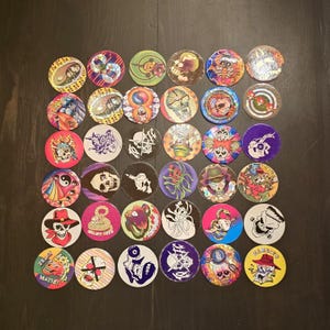 May include: A selection of colourful pogs featuring diverse designs, such as skulls and abstract patterns. The pogs are displayed on a dark surface, highlighting a range of colours and styles.