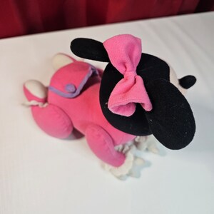 K&ouml;nnte beinhalten: Rosa Pl&uuml;schtier mit schwarzen Ohren und einer rosa Schleife. Das Spielzeug tr&auml;gt ein rosa-violettes Outfit mit wei&szlig;en Verzierungen.