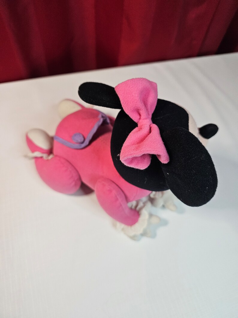 K&ouml;nnte beinhalten: Rosa Pl&uuml;schtier mit schwarzen Ohren und einer rosa Schleife. Das Spielzeug tr&auml;gt ein rosa-violettes Outfit mit wei&szlig;en Verzierungen.