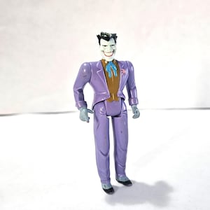 Puede incluir: Una figura de acción vintage del Joker, un villano de DC Comics. La figura viste un traje morado con chaleco marrón, pajarita azul y flor rosa. El Joker tiene guantes verdes y zapatos negros.