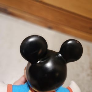 May include: A black Mickey Mouse figurine with large, round ears. The figurine is wearing a yellow shirt with a blue top and orange sleeves. The figure's white gloved hands are visible.