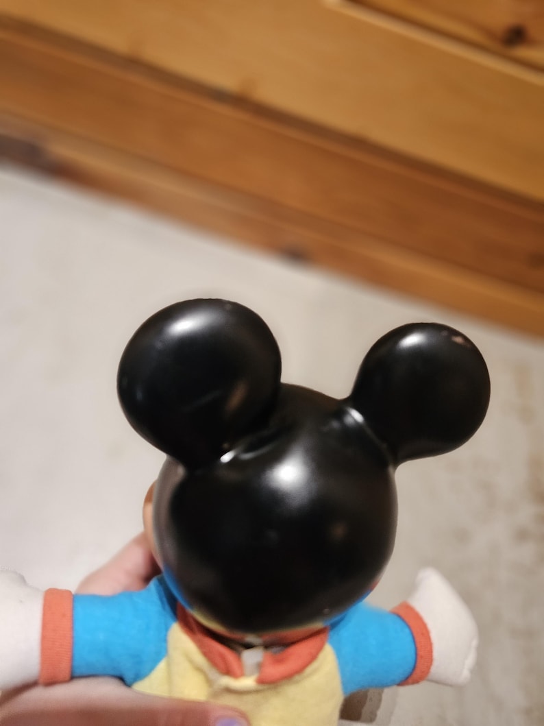 May include: A black Mickey Mouse figurine with large, round ears. The figurine is wearing a yellow shirt with a blue top and orange sleeves. The figure's white gloved hands are visible.