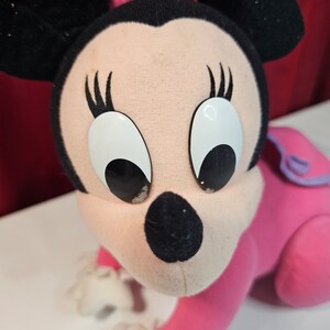 K&ouml;nnte beinhalten: Ein rosa und schwarzes Pl&uuml;schtier von Minnie Maus mit gro&szlig;en wei&szlig;en Augen und einer rosa Schleife. Das Spielzeug befindet sich in einer krabbelnden Position mit ausgestreckten Armen.