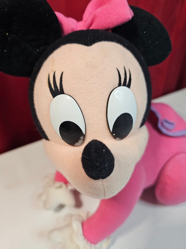 K&ouml;nnte beinhalten: Ein rosa und schwarzes Pl&uuml;schtier von Minnie Maus mit gro&szlig;en wei&szlig;en Augen und einer rosa Schleife. Das Spielzeug befindet sich in einer krabbelnden Position mit ausgestreckten Armen.