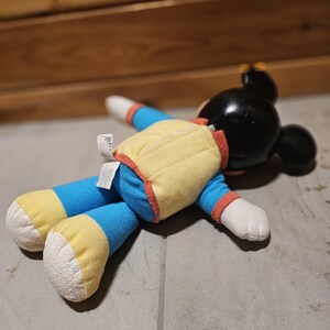 May include: A plush Mickey Mouse doll lying on a gray surface. The doll has a black head with large ears, a yellow vest with red trim, blue arms and legs with yellow cuffs, and white hands and feet.