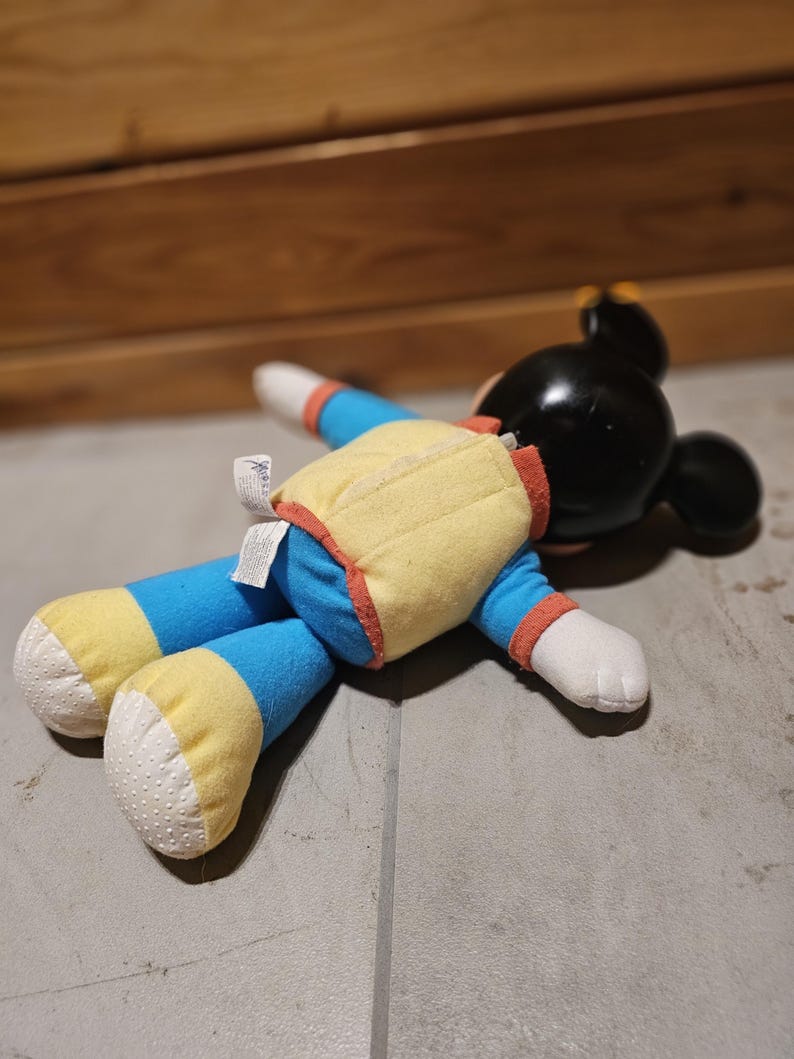 May include: A plush Mickey Mouse doll lying on a gray surface. The doll has a black head with large ears, a yellow vest with red trim, blue arms and legs with yellow cuffs, and white hands and feet.