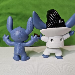 Peut inclure: Deux figurines Stitch bleues et blanches. Un Stitch porte un costume blanc et un chapeau noir. L'autre Stitch est bleu et a une couture blanche sur le dos.
