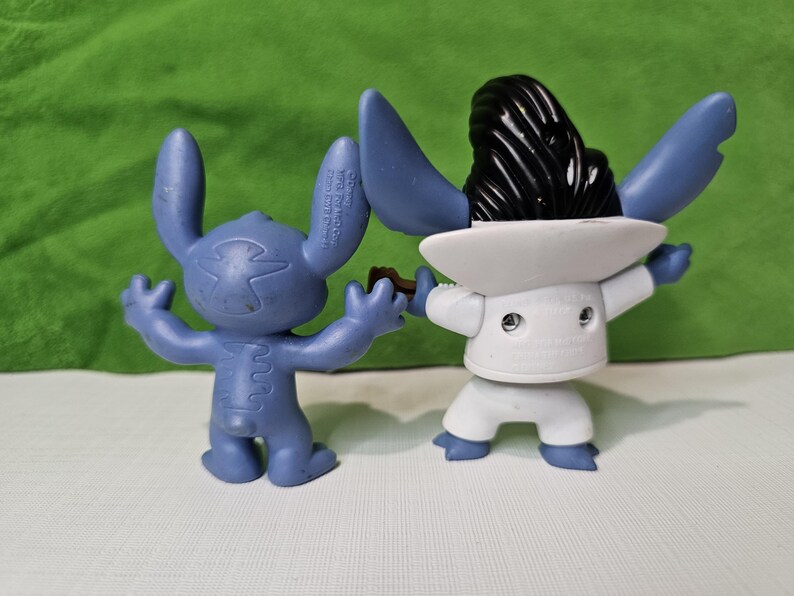 Peut inclure: Deux figurines Stitch bleues et blanches. Un Stitch porte un costume blanc et un chapeau noir. L'autre Stitch est bleu et a une couture blanche sur le dos.