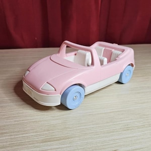 May include: A light pink toy convertible car with blue wheels and white accents. The car has a white interior with two seats and a windshield frame. The toy car is made of plastic.