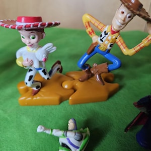 May include: A set of three plastic toy figures from the Toy Story franchise. The figures are Woody, Jessie, and Buzz Lightyear. Woody is wearing his signature cowboy hat and vest, Jessie is wearing her red and white cowgirl outfit, and Buzz Lightyear is wearing his space ranger suit. The figures are standing on a green surface.