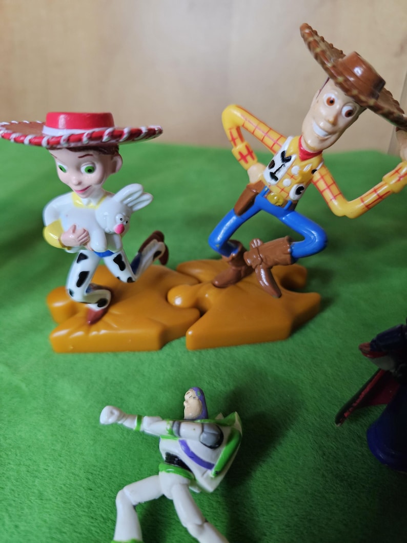 May include: A set of three plastic toy figures from the Toy Story franchise. The figures are Woody, Jessie, and Buzz Lightyear. Woody is wearing his signature cowboy hat and vest, Jessie is wearing her red and white cowgirl outfit, and Buzz Lightyear is wearing his space ranger suit. The figures are standing on a green surface.