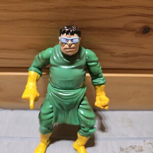 May include: A Dr. Octopus action figure. The figure is wearing a green outfit with yellow gloves and boots. The character has a stern expression and is wearing blue-tinted glasses.