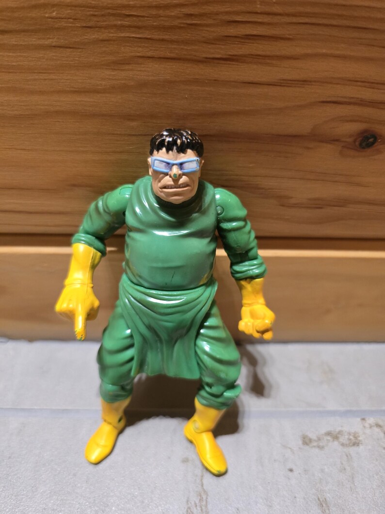 May include: A Dr. Octopus action figure. The figure is wearing a green outfit with yellow gloves and boots. The character has a stern expression and is wearing blue-tinted glasses.