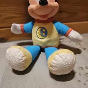 May include: A plush Mickey Mouse doll wearing a blue and yellow pajama set with a moon and stars emblem. The doll has black ears, a pink nose, and white gloves. The doll's feet are yellow with white dotted soles.