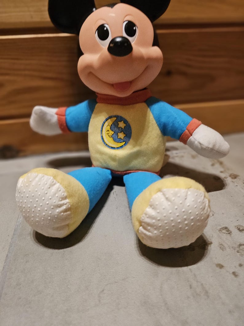 May include: A plush Mickey Mouse doll wearing a blue and yellow pajama set with a moon and stars emblem. The doll has black ears, a pink nose, and white gloves. The doll's feet are yellow with white dotted soles.