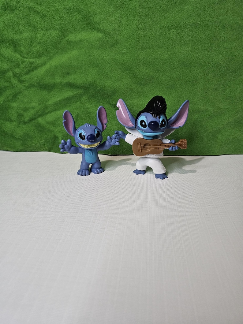 Peut inclure: Deux figurines bleues de Stitch du film Disney Lilo & Stitch. Un Stitch porte une combinaison blanche et tient une guitare brune. L'autre Stitch est debout la bouche ouverte.