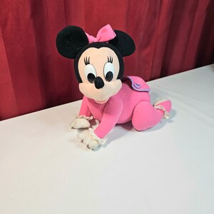 K&ouml;nnte beinhalten: Rosa Pl&uuml;schtier von Minnie Mouse, die ein rosa Kleid mit wei&szlig;er Spitzenborte und einer rosa Schleife tr&auml;gt. Sie krabbelt auf einer wei&szlig;en Oberfl&auml;che.