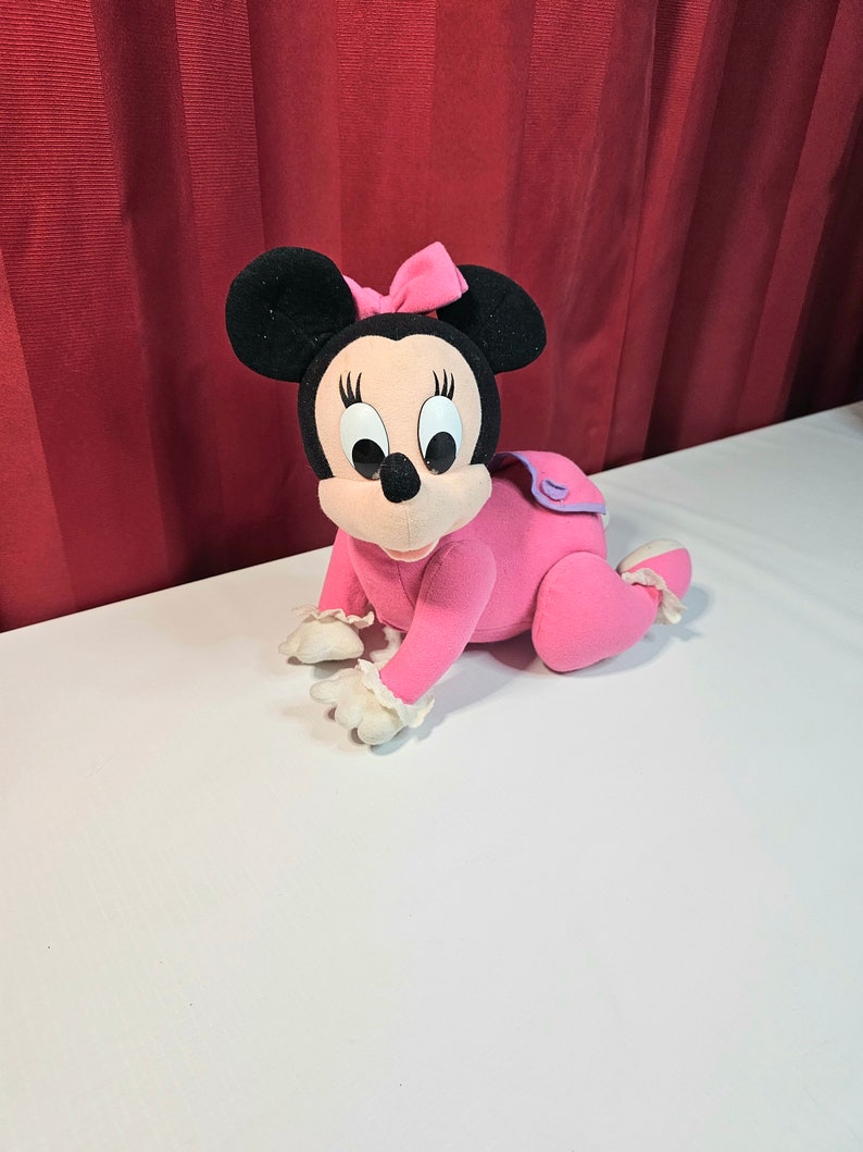 K&ouml;nnte beinhalten: Rosa Pl&uuml;schtier von Minnie Mouse, die ein rosa Kleid mit wei&szlig;er Spitzenborte und einer rosa Schleife tr&auml;gt. Sie krabbelt auf einer wei&szlig;en Oberfl&auml;che.