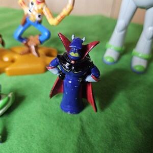 May include: A blue and red plastic toy figure with a black cape and a green face. The figure has horns and a black helmet. The figure is standing on a green surface.