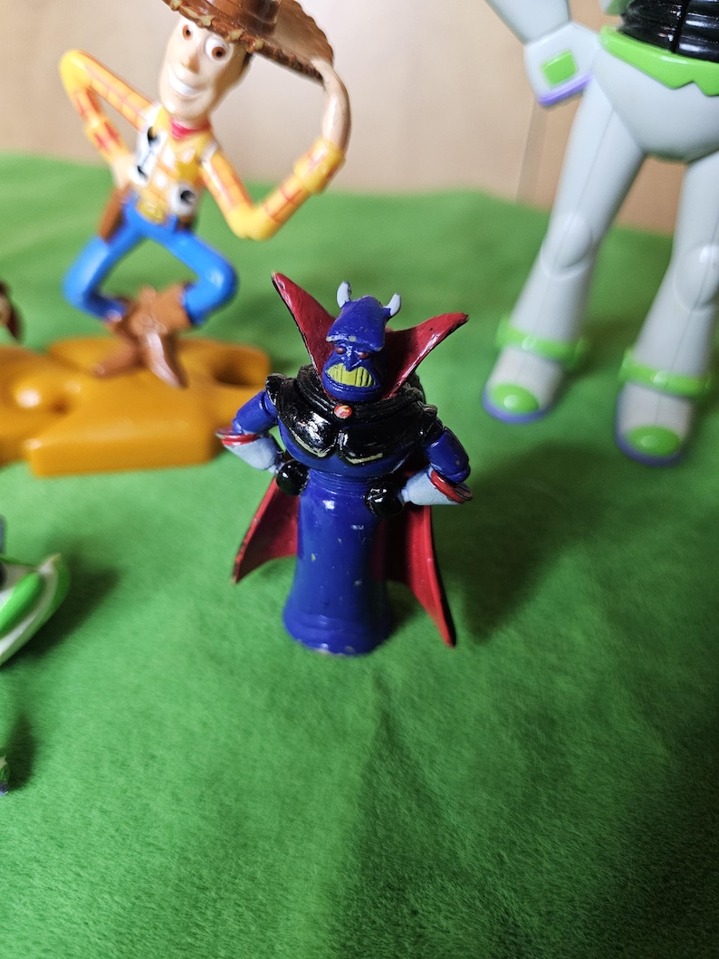 May include: A blue and red plastic toy figure with a black cape and a green face. The figure has horns and a black helmet. The figure is standing on a green surface.