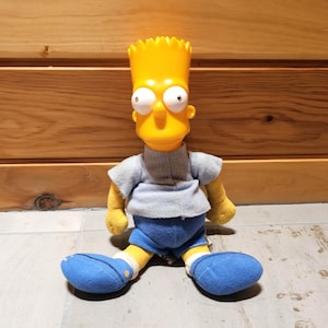 May include: A Bart Simpson plush doll with a yellow head and arms, wearing a gray shirt, blue shorts, and blue shoes. The doll has large, wide eyes and is sitting on a light-colored surface.