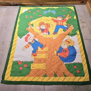 Peut inclure: Une courtepointe Cabbage Patch Kids représentant une maison dans les arbres avec quatre poupées. Les poupées portent des vêtements colorés et jouent dans la maison dans les arbres. La courtepointe est faite d'un tissu matelassé doux et a un schéma de couleurs vert, jaune et orange.