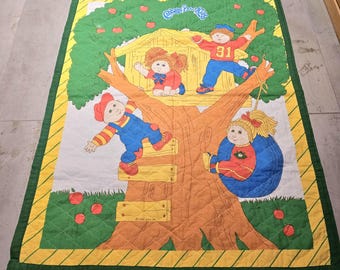 May include: A Cabbage Patch Kids quilt featuring a treehouse with four dolls. The dolls are wearing colorful clothing and are playing in the treehouse. The quilt is made of a soft, quilted fabric and has a green, yellow, and orange color scheme.