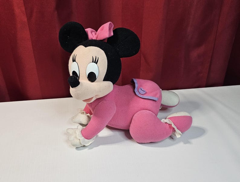 K&ouml;nnte beinhalten: Ein rosa und schwarzes Pl&uuml;schtier einer Comicmaus, die ein rosa Kleid und eine Schleife tr&auml;gt. Das Spielzeug krabbelt auf allen vieren.