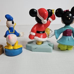 Op de afbeelding: Een set van vijf kleine plastic figuurtjes van populaire Disney-personages. De personages omvatten Mickey Mouse in een rood kerstmanpak, Minnie Mouse in een blauwe en paarse kimono, Donald Duck in een blauw matrozenpak, Woody uit Toy Story in een cowboy-outfit en een generieke Mickey Mouse in een gele en zwarte outfit.
