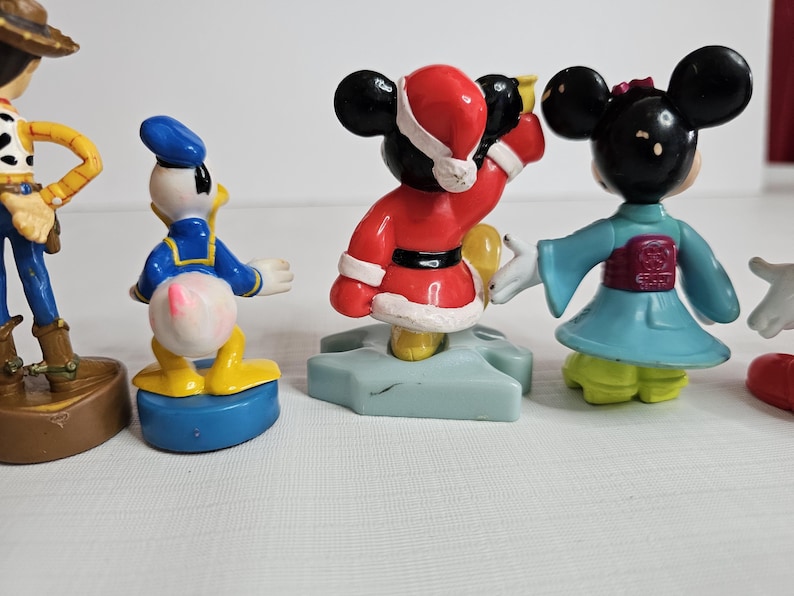 Op de afbeelding: Een set van vijf kleine plastic figuurtjes van populaire Disney-personages. De personages omvatten Mickey Mouse in een rood kerstmanpak, Minnie Mouse in een blauwe en paarse kimono, Donald Duck in een blauw matrozenpak, Woody uit Toy Story in een cowboy-outfit en een generieke Mickey Mouse in een gele en zwarte outfit.