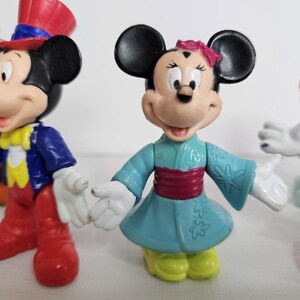 Op de afbeelding: Een set van vier plastic Disney-figuren. Mickey Mouse draagt een rood-blauwe hoge hoed en een blauw jasje. Minnie Mouse draagt een blauwe en witte kimono met een roze band.  Een andere Minnie Mouse draagt een paarse jurk met witte stippen en een rode strik. Goofy draagt een groene hoed en een bruin vest.
