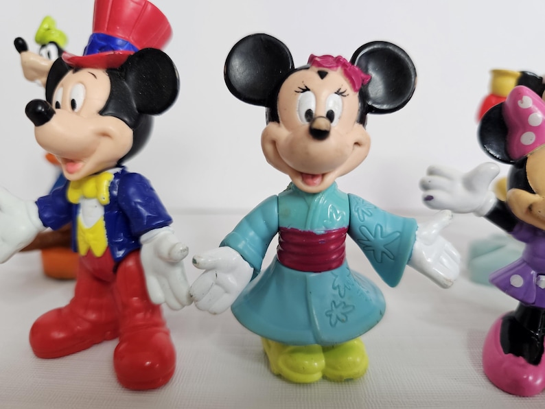 Op de afbeelding: Een set van vier plastic Disney-figuren. Mickey Mouse draagt een rood-blauwe hoge hoed en een blauw jasje. Minnie Mouse draagt een blauwe en witte kimono met een roze band.  Een andere Minnie Mouse draagt een paarse jurk met witte stippen en een rode strik. Goofy draagt een groene hoed en een bruin vest.