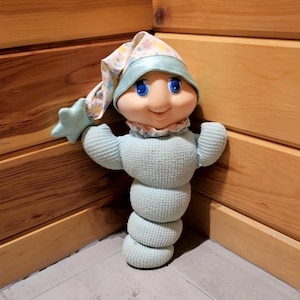 以下が含まれることがあります： 笑顔の顔、青い目、風変わりな帽子が特徴のヴィンテージぬいぐるみ。人形は、積み重ねられたデザインと腕を広げた薄い青色の体を持っています。帽子は星とドットのパターンでパステルカラーです。