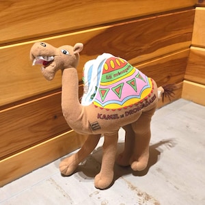 May include: A plush camel toy in shades of brown, with a colorful blanket draped over its back. The blanket features geometric patterns and text. The camel has a smiling expression and is standing on a light-colored surface.