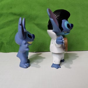 Peut inclure: Deux figurines bleues de Stitch du film Disney Lilo & Stitch. Un Stitch porte un smoking blanc et l'autre est dans une pose d&eacute;contract&eacute;e.