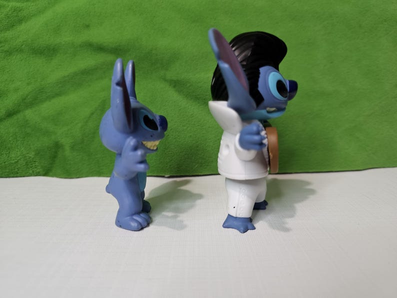 Peut inclure: Deux figurines bleues de Stitch du film Disney Lilo & Stitch. Un Stitch porte un smoking blanc et l'autre est dans une pose d&eacute;contract&eacute;e.