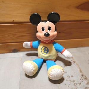 May include: A Mickey Mouse plush toy wearing a yellow and blue pajama set with a moon and stars graphic. The toy has black ears, a red collar, and white hands. The plush is sitting on a wooden surface.